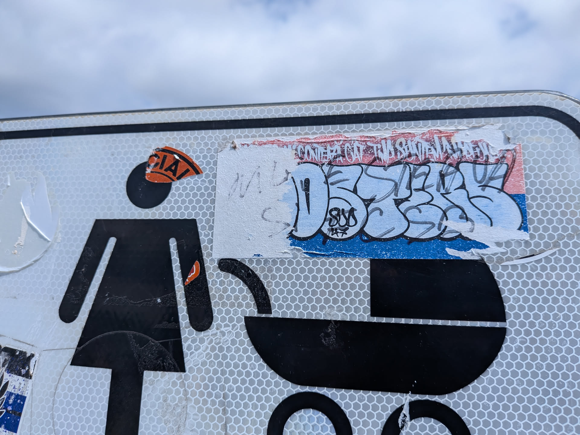 A torn rectangular sticker featuring light blue bubble graffiti text outlined in black over red, white, and blue stripes. The left portion is peeled away.