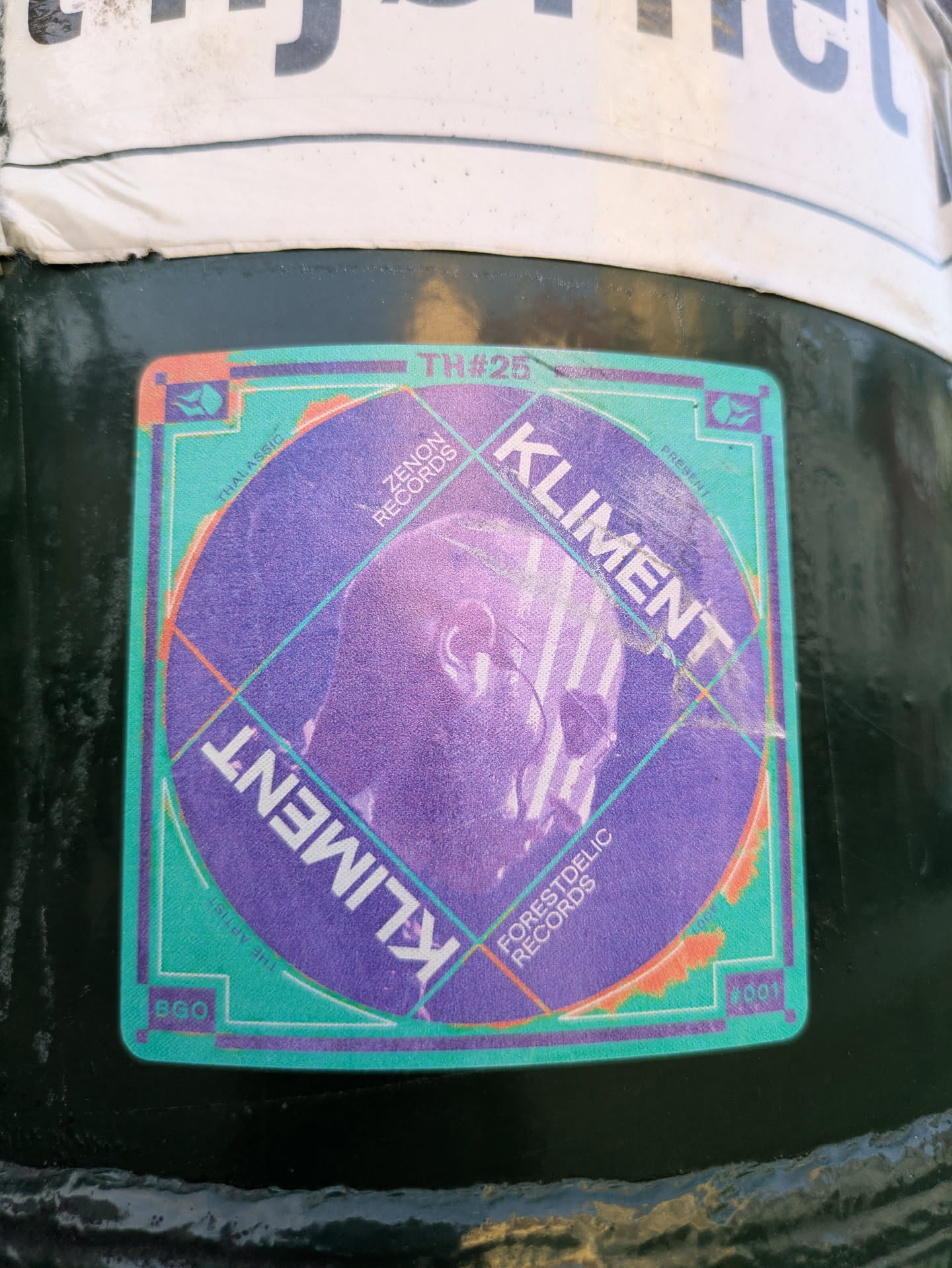The sticker is square with rounded corners, featuring a predominant color palette of purple, teal, and orange. The central element is a large purple circle containing a faint, stylized, almost ethereal profile of a head, possibly wearing glasses or a mask, overlaid with intersecting grid-like lines. The text 'KLIMENT' is prominently displayed twice, rotated, within this circle. Surrounding the circle is a teal border with purple and orange accents. Various record label names like 'ZENON RECORDS' and 'FORESTDELIC RECORDS' are visible within the design, along with 'THALASSIC' and event-related numbers like 'TH#25', 'BGO', and '001'. The overall design suggests a connection to electronic music or a record label promotion.