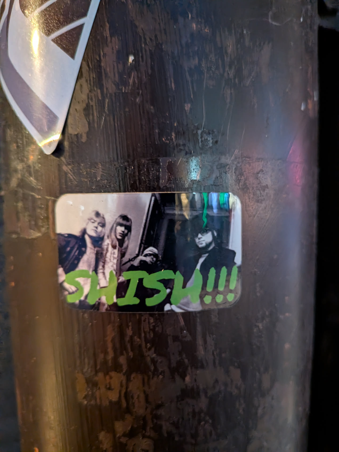 The sticker features a black and white photograph of three individuals, possibly a band, with a vibrant green text overlay. The individuals appear to be posing for the camera. The person on the left has blonde hair and is wearing a dark jacket. The person in the middle has dark hair and is wearing a light-colored, possibly sparkly, jacket. The person on the right is wearing a hat and sunglasses, with a dark jacket. The text 'SHISH!!!' is written in a bold, stylized green font across the bottom of the image, with exclamation points and dots adding emphasis. The sticker has rounded corners and appears to be slightly reflective, with hints of colorful light reflecting off its surface.