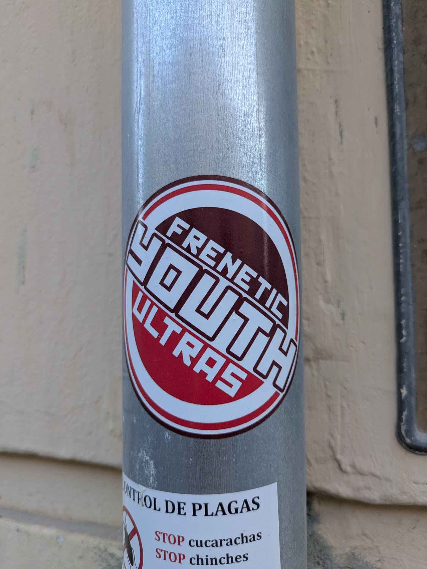 A round sticker with the words "Frenetic Youth Ultras" on a metal pole. The sticker has a white border, a red ring, and a maroon circle in the center. The words "Frenetic" and "Ultras" are in a red font, while the word "Youth" is in a white block font.