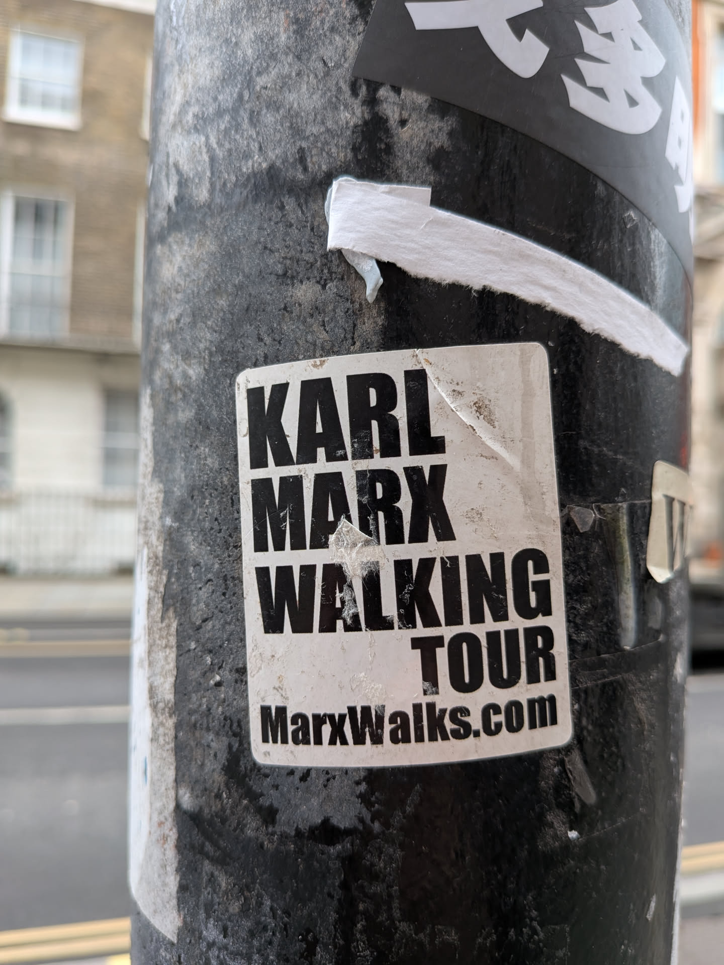 A rectangular sticker with slightly rounded corners, featuring bold black text on a white background. The text is arranged in a block format, reading 'KARL MARX' on the first line, 'WALKING' on the second, and 'TOUR' on the third. Below this, in a smaller font, is 'MarxWalks.com'. The sticker shows signs of wear, including dirt, scuff marks, and slight peeling at the edges, particularly the top right corner, indicating it has been exposed to the elements on a textured, dark pole.