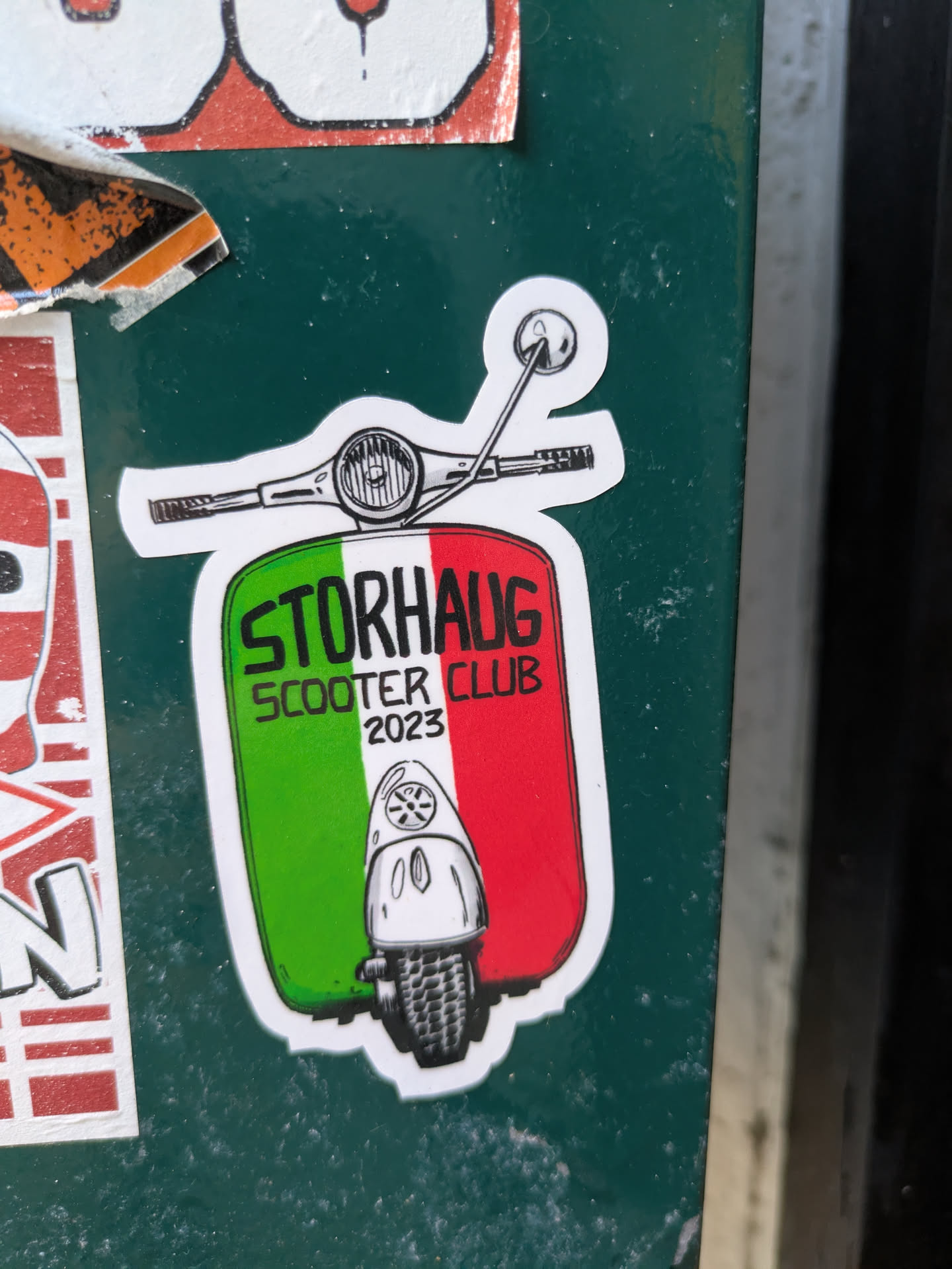 The sticker is shaped like the front view of a classic scooter, including handlebars and a front shield. The main body of the scooter is colored in the green, white, and red stripes of the Italian flag. Prominently displayed in white text is "STORHAUG SCOOTER CLUB" with "2023" written smaller below it. At the very bottom, on the red stripe, there's a small, stylized drawing of a vintage scooter's front wheel and fender.