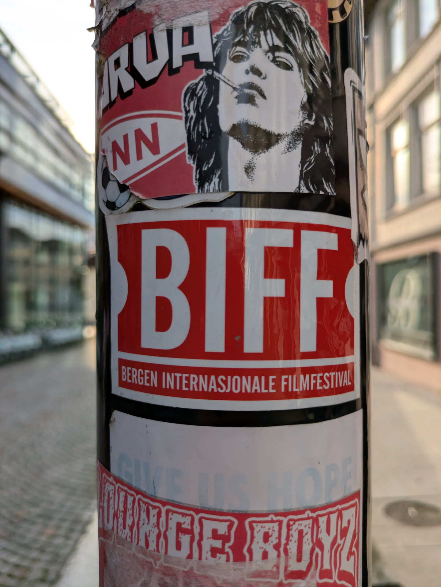 Street sticker The central focus of the image is a rectangular sticker on a dark pole. The sticker has a bold red background with 'BIFF' in large, white, sans-serif letters. Below this, in smaller white text, it reads 'BERGEN INTERNASJONALE FILMFESTIVAL'. Above this main sticker, a partially visible sticker features a black and white illustration of a person with long hair and a cigarette, along with fragmented text that includes 'ARUA' and 'NN' next to a small football icon. Below the BIFF sticker, another partially visible and faded sticker displays text that seems to read 'GIVE US HOPE' and 'OUNGE BOYZ' in a distressed font. The pole is situated outdoors, with a blurred cityscape in the background.