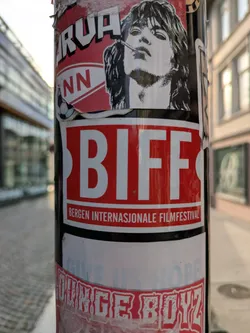 Street sticker The central focus of the image is a rectangular sticker on a dark pole. The sticker has a bold red background with 'BIFF' in large, white, sans-serif letters. Below this, in smaller white text, it reads 'BERGEN INTERNASJONALE FILMFESTIVAL'. Above this main sticker, a partially visible sticker features a black and white illustration of a person with long hair and a cigarette, along with fragmented text that includes 'ARUA' and 'NN' next to a small football icon. Below the BIFF sticker, another partially visible and faded sticker displays text that seems to read 'GIVE US HOPE' and 'OUNGE BOYZ' in a distressed font. The pole is situated outdoors, with a blurred cityscape in the background.