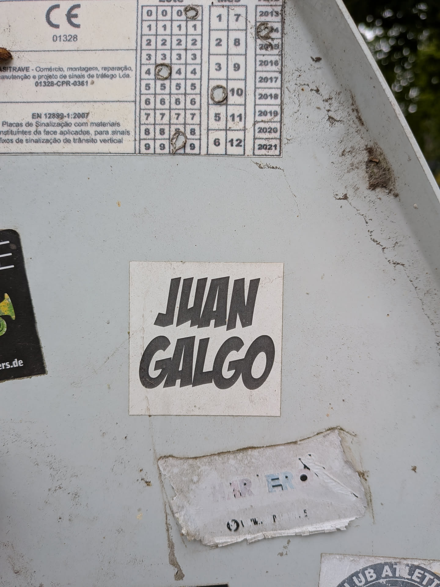 A square white sticker featuring the name 'JUAN GALGO' in bold, stylized black lettering with a slight slant, affixed to a grey metal surface.