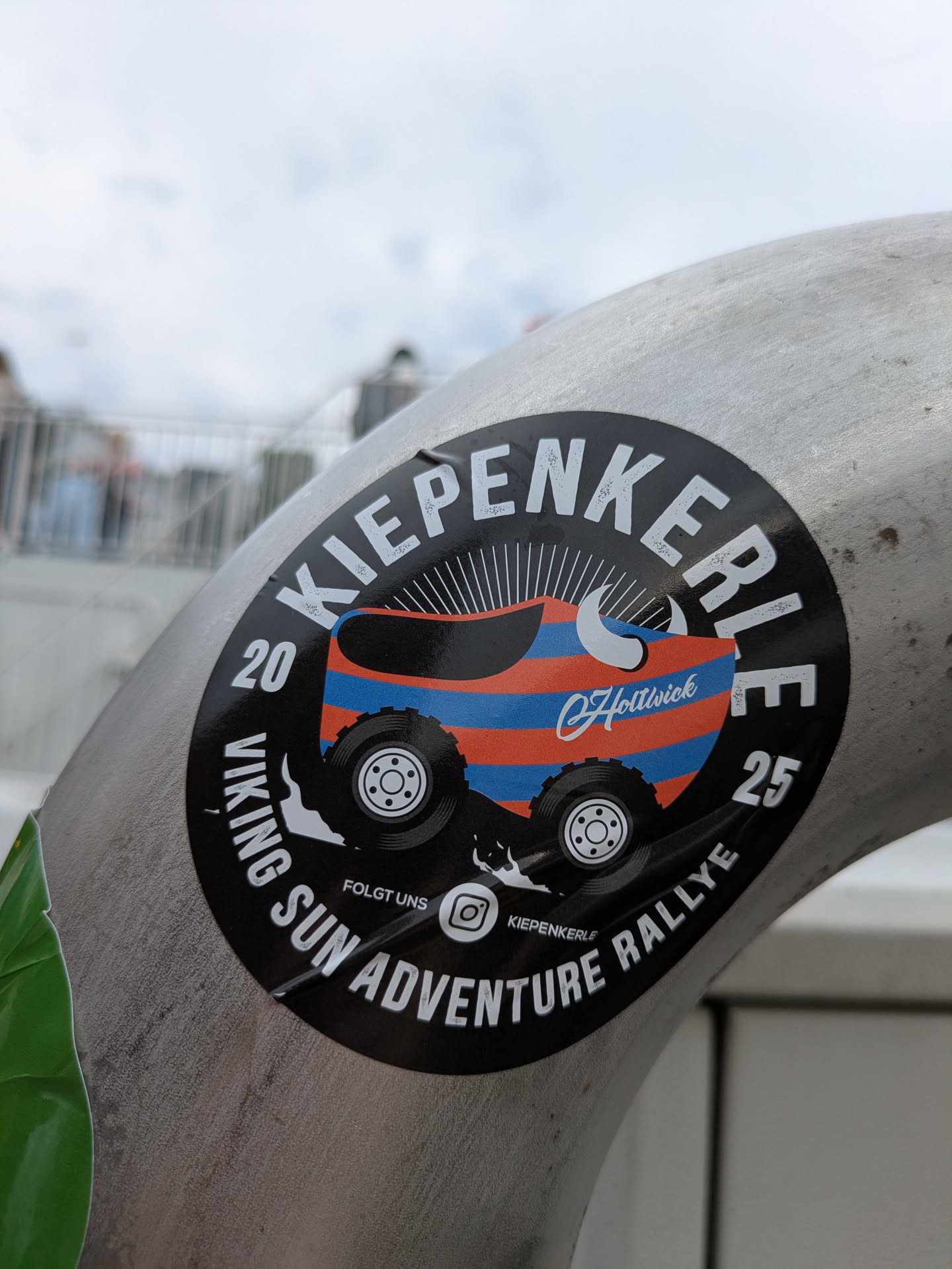 Street sticker This is a round, black sticker applied to a metal pipe. The sticker's central design features a highly stylized, colorful vehicle resembling a traditional German wooden clog (Kiepe), painted with prominent orange and blue horizontal stripes. This 'clog-car' is depicted with large, rugged off-road tires. A white Viking-style horn or tusk protrudes from the front of the vehicle, and sun rays emanate from behind it, suggesting 'Sun' in the rally name. The outer edge of the sticker contains white text. The bottom states 'VIKING SUN ADVENTURE RALLYE' in an arc, flanked by '20' on the left and '25' on the right, likely indicating years. At the top, in an arc, is the word 'KIEPENKERLE'. Below the main graphic, there's a small Instagram logo with 'FOLGT UNS' (Follow Us) and '@KIEPENKERLE' next to it. A small copyright symbol followed by 'Hollwick' is visible on the vehicle itself.
