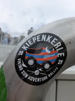Street sticker This is a round, black sticker applied to a metal pipe. The sticker's central design features a highly stylized, colorful vehicle resembling a traditional German wooden clog (Kiepe), painted with prominent orange and blue horizontal stripes. This 'clog-car' is depicted with large, rugged off-road tires. A white Viking-style horn or tusk protrudes from the front of the vehicle, and sun rays emanate from behind it, suggesting 'Sun' in the rally name. The outer edge of the sticker contains white text. The bottom states 'VIKING SUN ADVENTURE RALLYE' in an arc, flanked by '20' on the left and '25' on the right, likely indicating years. At the top, in an arc, is the word 'KIEPENKERLE'. Below the main graphic, there's a small Instagram logo with 'FOLGT UNS' (Follow Us) and '@KIEPENKERLE' next to it. A small copyright symbol followed by 'Hollwick' is visible on the vehicle itself.