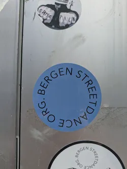 Street sticker The sticker is a perfectly circular shape with a light blue background. Black capital letters are arranged in a circle around the perimeter, reading 'ORG. BERGEN STREETDANCE'. The text is sans-serif and curves along the edge, with 'ORG. BERGEN' at the top and 'STREETDANCE' at the bottom. The center of the sticker is left blank.