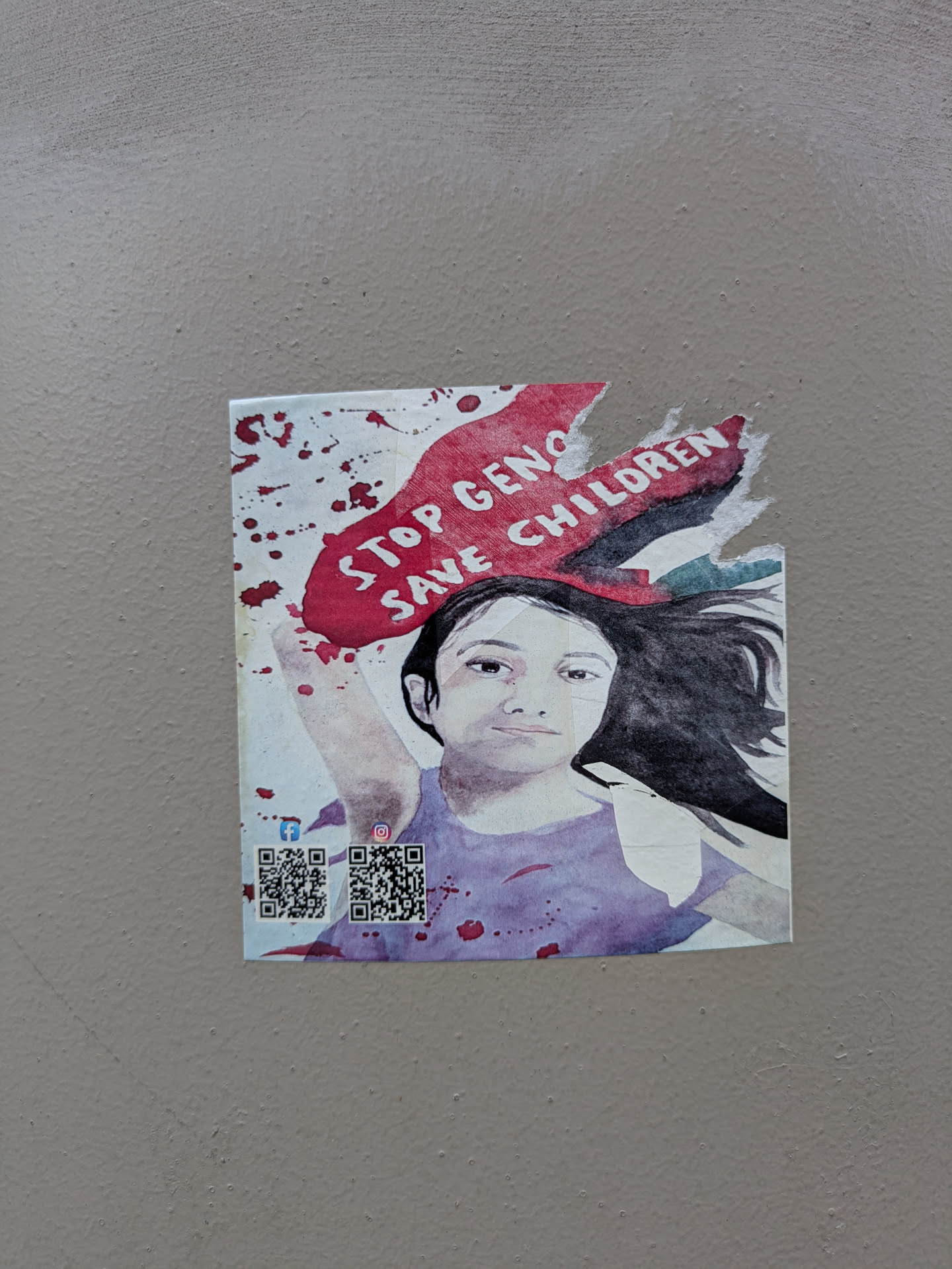 The sticker is pasted on a plain, light-colored wall. It features a stylized illustration of a child with dark hair and a purple shirt, looking directly at the viewer. Above the child's head, there's a large, torn red shape, resembling a banner or a torn flag, with the white text "STOP GENOCIDE SAVE CHILDREN" written across it. The background is white, splattered with numerous red droplets, resembling blood. The sticker shows signs of wear, with some tearing, particularly at the top right edge of the red shape. In the bottom left corner, there are two distinct QR codes. Above the left QR code is the Facebook 'f' logo, and above the right QR code is the Instagram camera logo, indicating links to social media platforms.