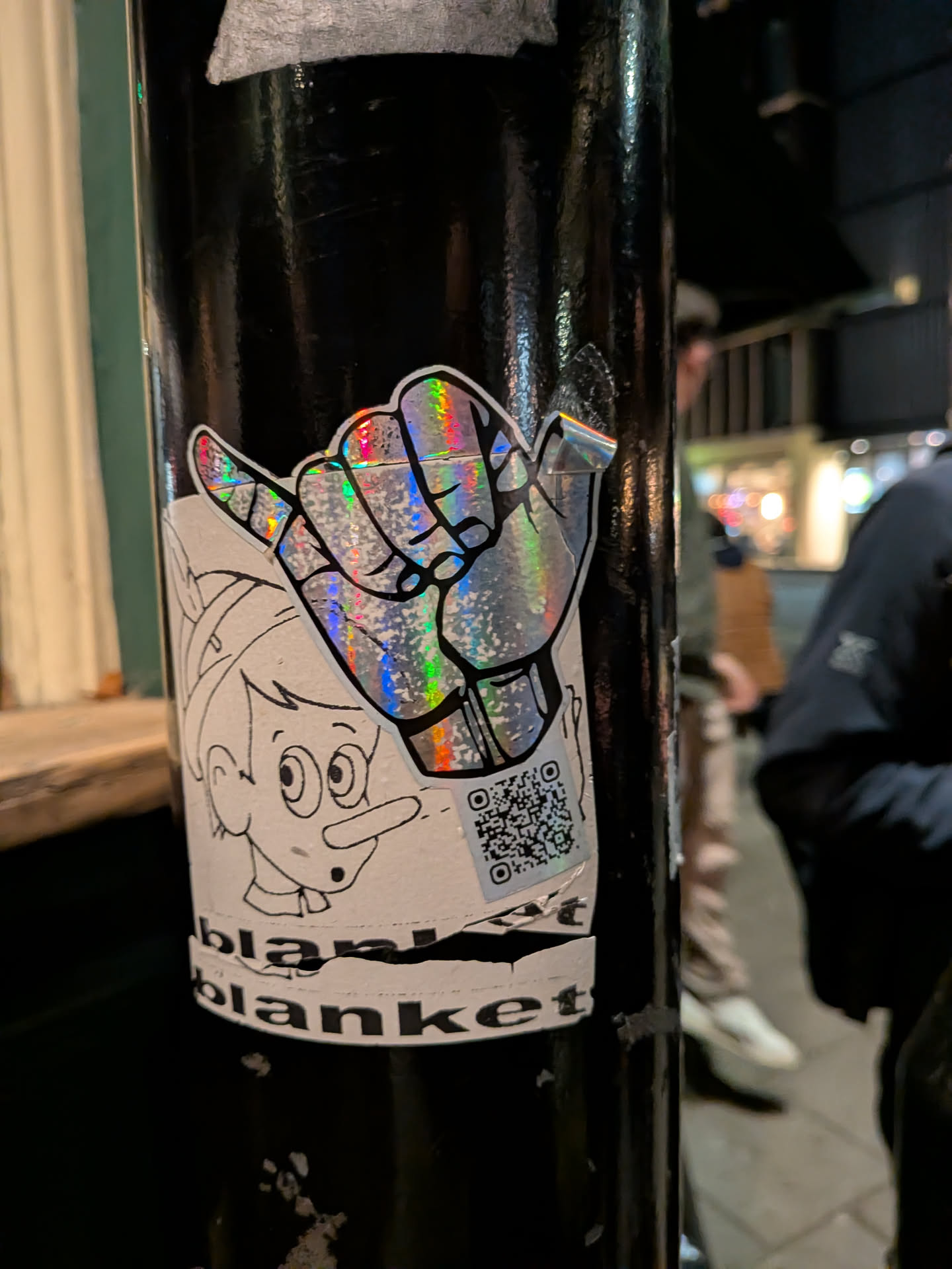 A white sticker with black outlines featuring two main elements: a large, stylized hand making a 'shaka' (thumb and pinky extended) gesture, and a cartoon character's head resembling Pinocchio with a long nose, positioned below and to the left of the hand. The hand is filled with a highly reflective, holographic material that displays a rainbow spectrum of colors (greens, blues, reds, yellows) due to light reflection. The Pinocchio-like character is drawn in simple black outlines on a white background. To the right of the cartoon character and below the holographic hand, there is a black and white QR code. Below the cartoon character, the word 'blanket' is visible in black sans-serif font, with some tearing at the top edge. The sticker is applied to a black pole, showing some minor wear and slightly peeling edges.