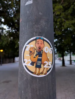 A round sticker is affixed to a pole. The sticker features a cartoonish camel with a human face and sunglasses. The camel wears a blue scarf with the words "COMO 1907". Around the edge of the sticker are numbers.