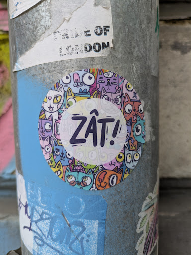 This is a round sticker affixed to a pole. The sticker is densely packed with cartoon cats of various colors and expressions. In the center, the word "ZÂT!" is written in bold, purple letters.