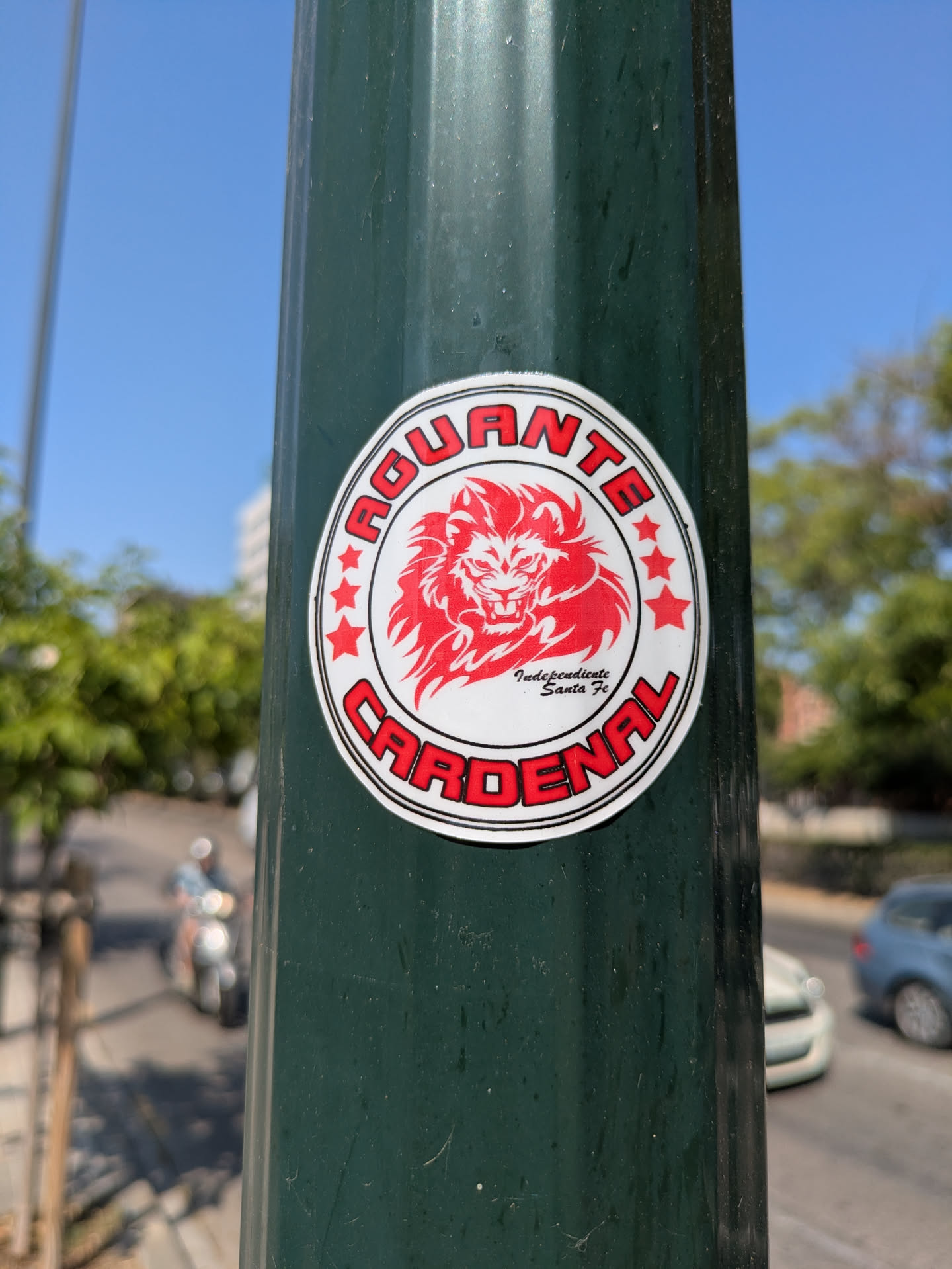 Street sticker A round sticker is affixed to a green pole. The sticker has a white background with a red border. Around the top border of the sticker, in red letters, is the word "AGUANTE." At the bottom border of the sticker is the word "CARDENAL," also in red letters. In the center of the sticker is a stylized red lion's head. Below the lion's head is the text "Independiente Santa Fe". There are also five red stars along the side.