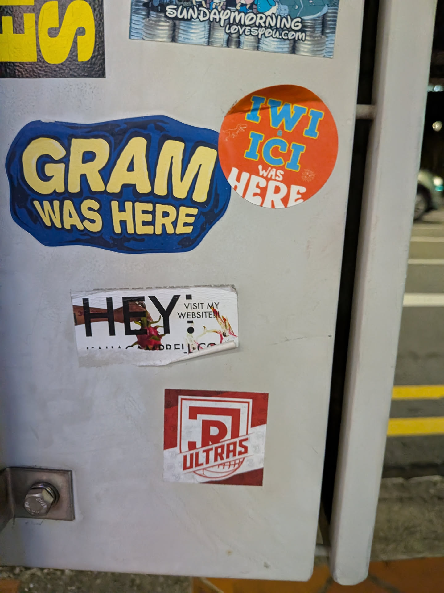 A rectangular white sticker featuring the bold word 'HEY' integrated with an image of a hand holding a flower. It includes a call to action to visit a website, with the URL partially obscured by peeling.