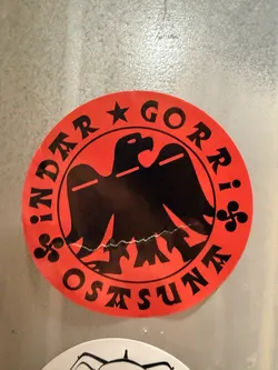 Street sticker A circular sticker with a bright red background and black elements. At its center is a stylized black eagle, facing left with its wings spread. The eagle has a sharp beak and a single eye, and its lower body features a jagged, wave-like or mountainous outline. Encircling the eagle is black text: 'INDER ★ GORRI' along the top arc and 'OSTASUNA' along the bottom arc. A black five-pointed star separates 'INDER' and 'GORRI'. Two black Lauburu symbols (a traditional Basque cross with curved arms) are positioned on either side of the eagle, separating 'GORRI' from 'OSTASUNA'. The sticker shows minor signs of wear, including a slight crease or imperfection near the bottom edge of the eagle.