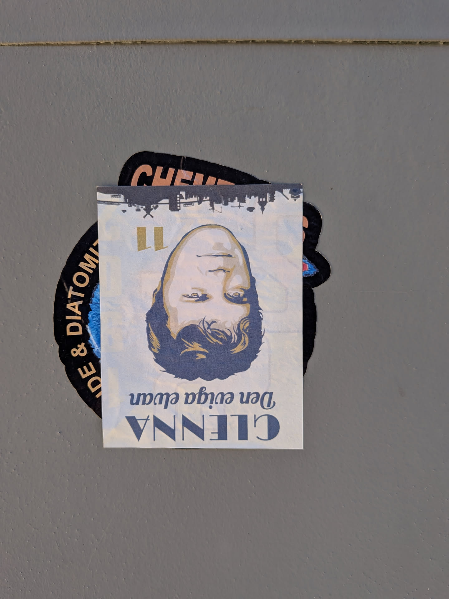 The sticker is a rectangular piece of paper with a portrait of a person's face, oriented upside down. The face is rendered in a stylized, illustrative manner with bold lines and a limited color palette of skin tones, dark blue for hair and shadows, and yellow for accents. Above the face, a silhouette of a cityscape is depicted. Below the face, text is printed in a vintage-style font. The sticker is partially overlapping a larger, black, circular sticker with white text and a blue fuzzy center.
