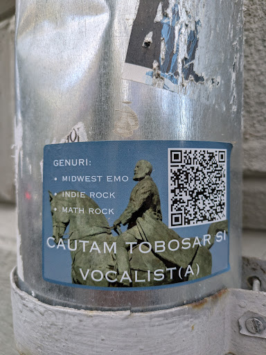 A blue sticker featuring an image of a man on a horse is adhered to a metallic pole. The sticker lists musical genres such as Midwest Emo, Indie Rock, and Math Rock. Below the image, text indicates the band is searching for a drummer and a vocalist. A QR code is located in the upper right corner of the sticker.
