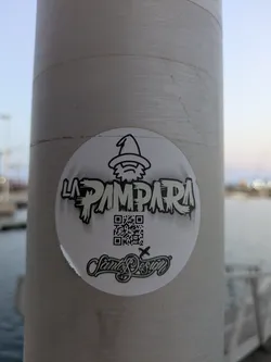 Street sticker A round white sticker of the band La Pampara is stuck on a grey pole. The sticker features a logo of a wizard above the band name, a QR code, and the name Santos Design.