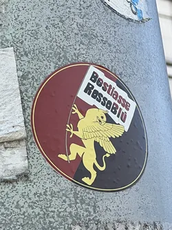 Street sticker A circular sticker featuring a golden winged lion standing on its hind legs, holding a white flag. The background is split vertically, with the left half in dark red/maroon and the right half in dark blue/black. A thin golden-yellow border encircles the design. The lion has a fierce expression and prominent wings. The flag it holds displays the text 'Bestiasse' in black and 'RossoBlü' in red.