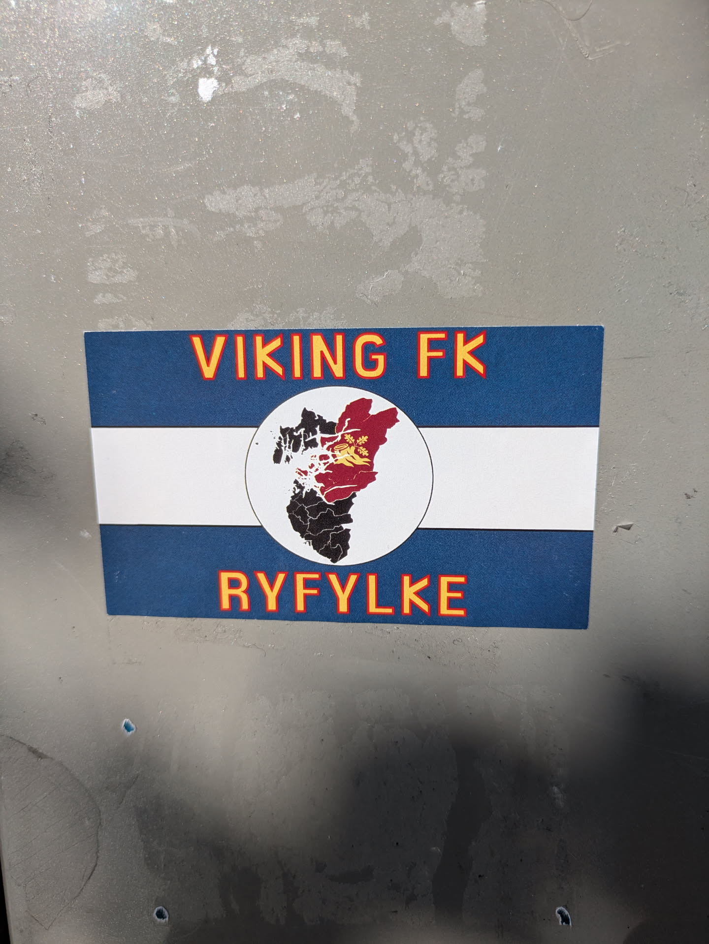 The sticker is rectangular with a design resembling a flag, featuring a wide blue band at the top, a white horizontal stripe in the middle, and another wide blue band at the bottom. In the center of the white stripe, there's a circular emblem. This emblem contains a stylized map or geographic outline, with parts colored red and black, and a yellow symbol (possibly a crown or emblem) on the red section. Above the central emblem, the text 'VIKING FK' is written in orange-yellow letters with a red outline. Below the emblem, 'RYFYLKE' is written in the same style. The overall design suggests support for the football club Viking FK and its connection to the Ryfylke region.