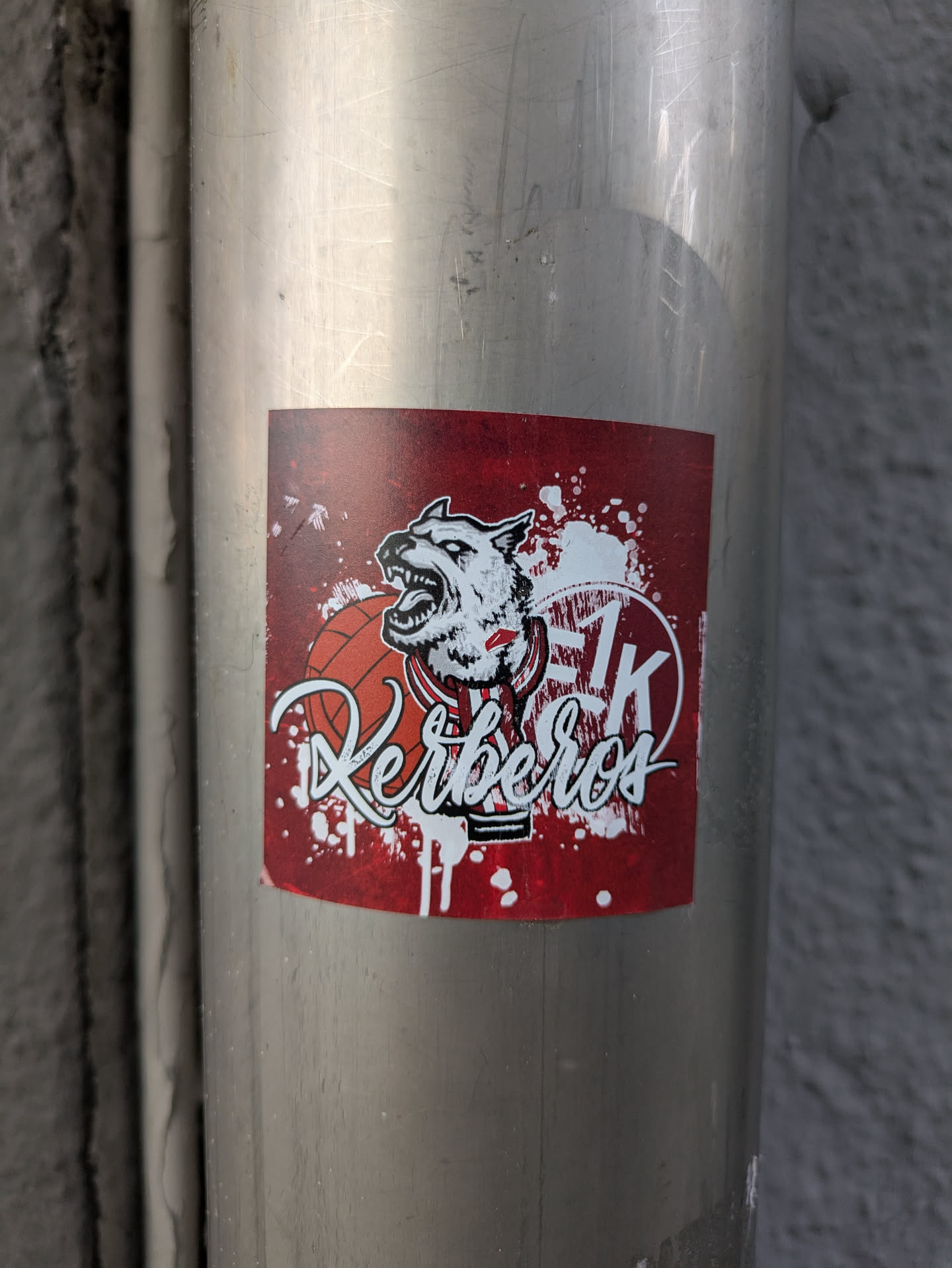 The sticker features a stylized, aggressive white canine head with an open mouth and sharp teeth, wearing a red and white striped collar. A basketball is partially visible to the left of the head. Below these elements, the word 'Kerberos' is written in a dynamic white script font. To the upper right, the initials 'SK' are prominently displayed in white within a partial circular outline. The entire design is set against a dark red background with white paint splatter effects, giving it a rough, energetic aesthetic. The sticker is placed on a silver metal pipe.