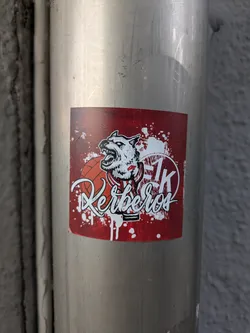 Street sticker The sticker features a stylized, aggressive white canine head with an open mouth and sharp teeth, wearing a red and white striped collar. A basketball is partially visible to the left of the head. Below these elements, the word 'Kerberos' is written in a dynamic white script font. To the upper right, the initials 'SK' are prominently displayed in white within a partial circular outline. The entire design is set against a dark red background with white paint splatter effects, giving it a rough, energetic aesthetic. The sticker is placed on a silver metal pipe.