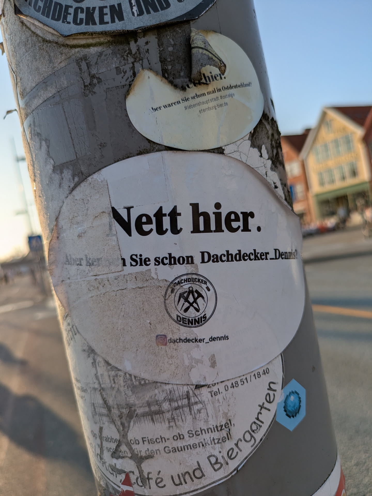 A prominent white, oval-shaped sticker is attached to a weathered, gray utility pole. The sticker features bold black text at the top, 'Nett hier.' (Nice here.). Below this, it continues with 'Aber kennen Sie schon Dachdecker_Dennis?' (But do you already know Roofer_Dennis?), though parts of this text are slightly obscured. Central to the sticker is a black circular logo with crossed roofer's hammers and the words 'DACHDECKER DENNIS'. Below the logo, an Instagram handle is visible. The sticker exhibits signs of wear, with some peeling edges and dirt, and is partially layered over other older stickers.