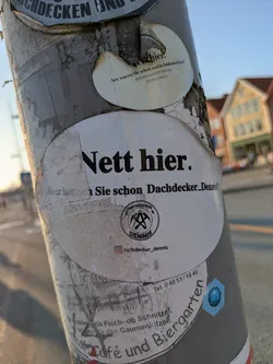 Street sticker A prominent white, oval-shaped sticker is attached to a weathered, gray utility pole. The sticker features bold black text at the top, 'Nett hier.' (Nice here.). Below this, it continues with 'Aber kennen Sie schon Dachdecker_Dennis?' (But do you already know Roofer_Dennis?), though parts of this text are slightly obscured. Central to the sticker is a black circular logo with crossed roofer's hammers and the words 'DACHDECKER DENNIS'. Below the logo, an Instagram handle is visible. The sticker exhibits signs of wear, with some peeling edges and dirt, and is partially layered over other older stickers.