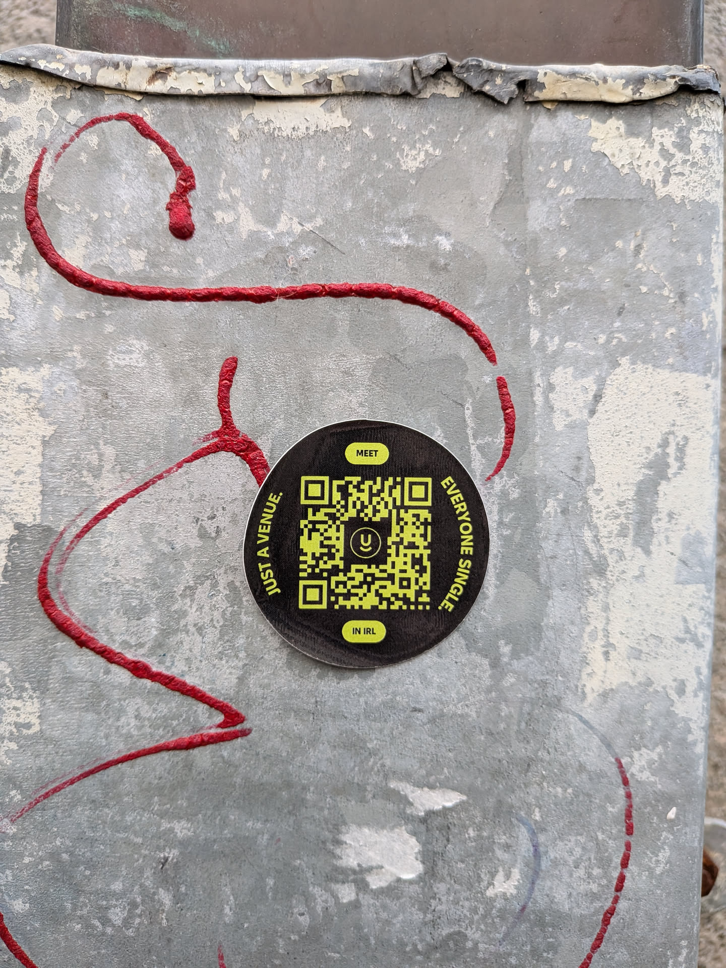 A circular sticker with a black background. In the center is a bright yellow QR code with a stylized 'U' logo in the middle. Around the QR code, text is arranged in a circle. The top reads 'MEET' in a yellow rectangle. The left side reads 'JUST A VENUE.' in white text. The right side reads 'EVERYONE SINGLE.' in white text. The bottom reads 'IN IRL' in a yellow rectangle.