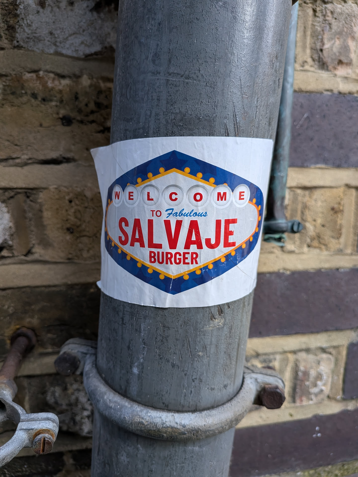 The sticker is an oval-shaped design, mimicking the iconic 'Welcome to Fabulous Las Vegas' sign. It features a dark blue border with white star-like accents and is outlined by a string of yellow-gold circular lights. The main background is white. At the top, the word 'WELCOME' is spelled out in individual white circles with red letters. Below this, in a smaller, cursive-like blue font, it reads 'TO Fabulous'. The central and largest text is 'SALVAJE' in bold red capital letters, followed by 'BURGER' in smaller yellow-gold capital letters. The sticker is applied to a grey metal pipe and shows slight wear and peeling at its edges.