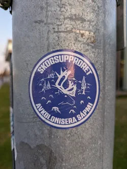Street sticker A round blue sticker with a white outline featuring a reindeer and text in Swedish. The text on the sticker translates to "Forest Uprising: Decolonize Sápmi".