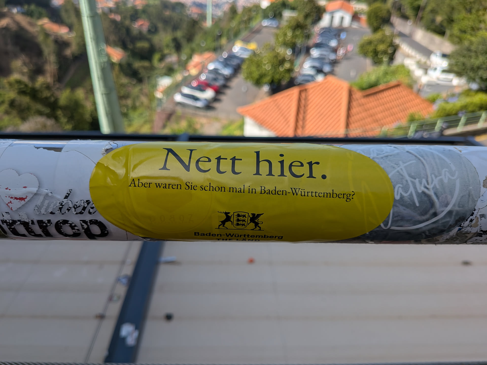 An oval yellow sticker on a metal bar. It displays the German text 'Nett hier. Aber waren Sie schon mal in Baden-Württemberg?' alongside the state's coat of arms and 'Baden-Württemberg THE LÄND' branding.