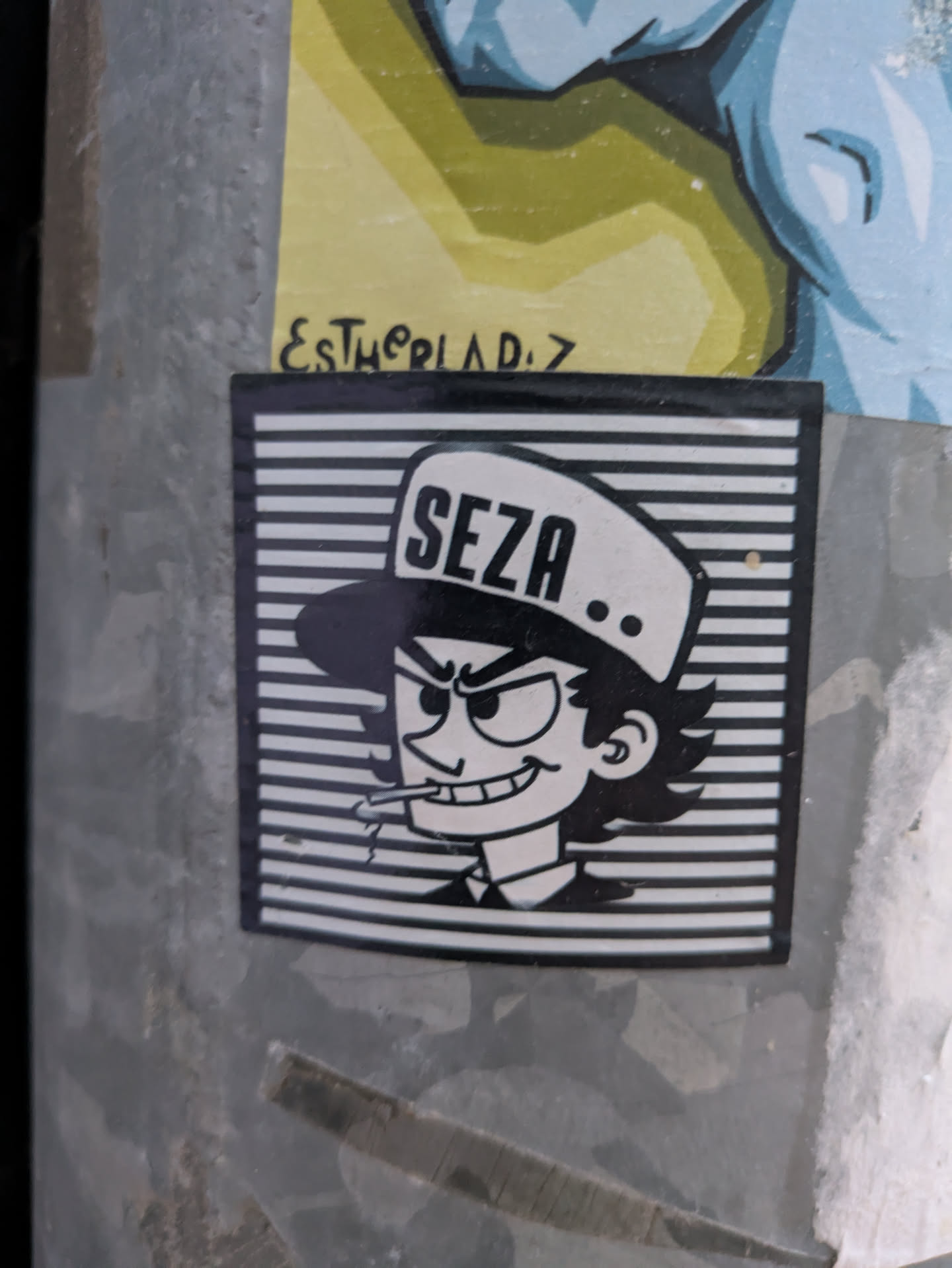 This black and white sticker features a cartoon character with a hat labeled "SEZA". The character has a mischievous smirk and is smoking a cigarette. The background is composed of horizontal black and white lines. The sticker is on a metallic surface.