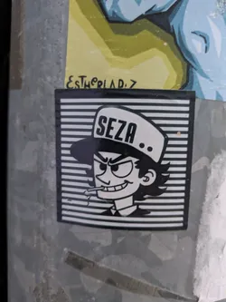 Street sticker This black and white sticker features a cartoon character with a hat labeled "SEZA". The character has a mischievous smirk and is smoking a cigarette. The background is composed of horizontal black and white lines. The sticker is on a metallic surface.
