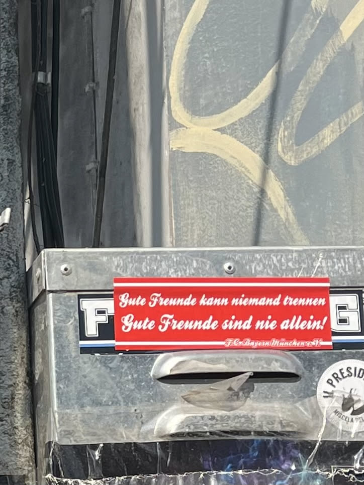 A rectangular sticker with a bright red background and white text. The main text is in a decorative, cursive-like font, while the club name at the bottom is in a smaller, sans-serif font. The sticker appears slightly worn and adhered to a metallic surface, possibly a utility box or pole.