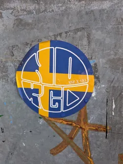 Street sticker The sticker is circular and predominantly features the colors blue and yellow, arranged in a pattern that evokes the Swedish flag. A thick white outline encircles the sticker. Within the blue and yellow fields, a white graphic design is overlaid. This design consists of abstract, angular shapes and lines, forming what appears to be stylized lettering or a logo. In the upper right quadrant of the sticker, within a yellow band, the text 'NSF X SKI' is visible in small, white, sans-serif font. The overall impression is modern and graphic.