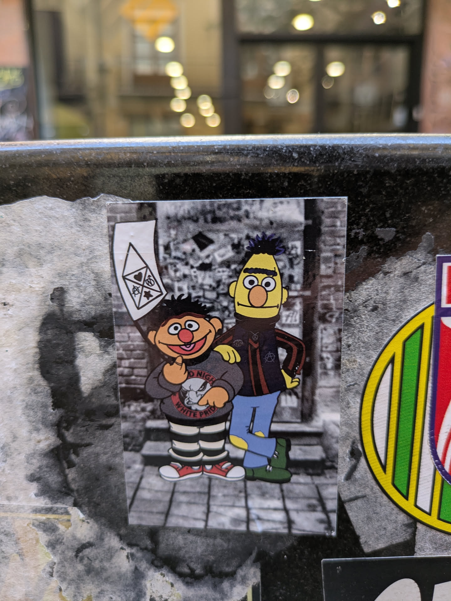 This sticker shows Ernie and Bert edited to have a punk look, on top of a black and white crowd background. Ernie is on the left holding a flag with some symbols, wearing striped pants and a sweater that reads "Good Night, White Pride" and an image of a cartoonish bulldog. Bert on the right wears a leather jacket with a symbol of anarchy.