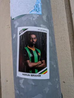 Street sticker The sticker features a portrait of a man, presumably a footballer, named Harun Ibrahim. He is wearing a green and black striped jersey with a yellow and red trim around the collar. The jersey has a 'SELECT' logo and a crest on the left chest. The man has his arms crossed and is looking directly at the camera with a serious expression. The background is dark, creating a dramatic effect. The sticker has a white border with a green and black geometric design in the top left corner and a stripe of green, yellow, and red at the bottom. The text 'ATLETETERNA' is visible at the top, and 'HARUN IBRAHIM' is at the bottom.