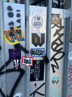 Street sticker The sticker at the center of the image is a circular sticker with a white border. Inside, it depicts a stylized atom with a purple nucleus and blue electron paths. The word "ELEKTRON" is written in white capital letters above the atom. Below the atom, smaller text reads "SOCIAL SPHERE ART SES". There is also an Instagram handle "@elektron.events" in white text at the bottom of the sticker. The background of this sticker is dark purple. To its left is a yellow and red sticker with the word "CHILL" in stylized blue and yellow lettering with a red outline. To the right of the 'Elektron' sticker is a square black sticker with white cartoonish rabbit ears and a red ribbon tied around the body of a white figure with black accents.