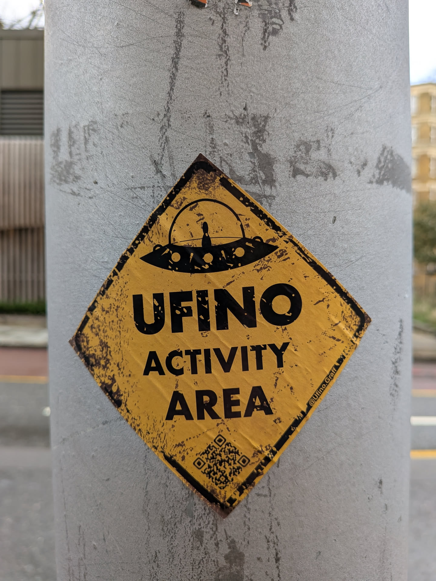 The sticker is diamond-shaped, resembling a weathered warning sign. It features a distressed yellow background with bold black text and imagery. At the top, a black silhouette of a classic flying saucer (UFO) is depicted, with a small figure visible inside its dome. Below the UFO, the prominent text reads "UFINO" followed by "ACTIVITY AREA". The entire sticker has a grunge aesthetic, with black splotches and faded areas giving it an aged, worn appearance. A social media handle is visible in the bottom right, and a QR code is present in the bottom left.