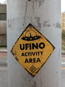 Street sticker The sticker is diamond-shaped, resembling a weathered warning sign. It features a distressed yellow background with bold black text and imagery. At the top, a black silhouette of a classic flying saucer (UFO) is depicted, with a small figure visible inside its dome. Below the UFO, the prominent text reads "UFINO" followed by "ACTIVITY AREA". The entire sticker has a grunge aesthetic, with black splotches and faded areas giving it an aged, worn appearance. A social media handle is visible in the bottom right, and a QR code is present in the bottom left.