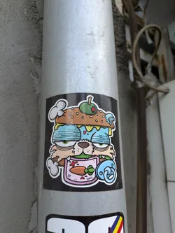 Street sticker The sticker features a cartoon cat with a burger-like head, complete with a burger bun top, an olive, and mushrooms. The cat has drowsy eyes and a bib with a carrot printed on it. A pacifier hangs nearby. The sticker's design is quirky and somewhat surreal.