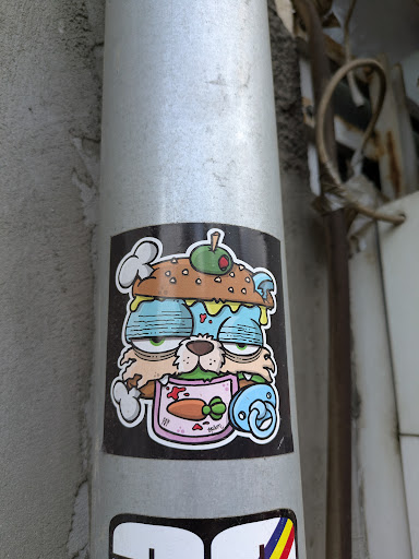 The sticker features a cartoon cat with a burger-like head, complete with a burger bun top, an olive, and mushrooms. The cat has drowsy eyes and a bib with a carrot printed on it. A pacifier hangs nearby. The sticker's design is quirky and somewhat surreal.