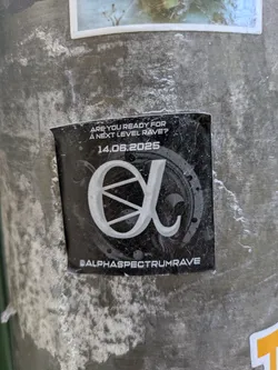 Street sticker A black sticker advertising an upcoming event. It features the text "ARE YOU READY FOR A NEXT LEVEL RAVE?" and the date "14.06.2025" above a stylized logo resembling the Greek letter alpha within a speaker graphic. At the bottom, the social media handle @ALPHASPECTRUMRAVE is printed. The sticker is affixed to a weathered metal pole.