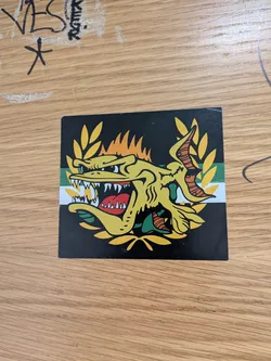 Street sticker A square sticker featuring a stylized, aggressive yellow fish with sharp teeth and an open mouth. The fish is framed by a yellow laurel wreath against a black background with a horizontal green and white stripe.