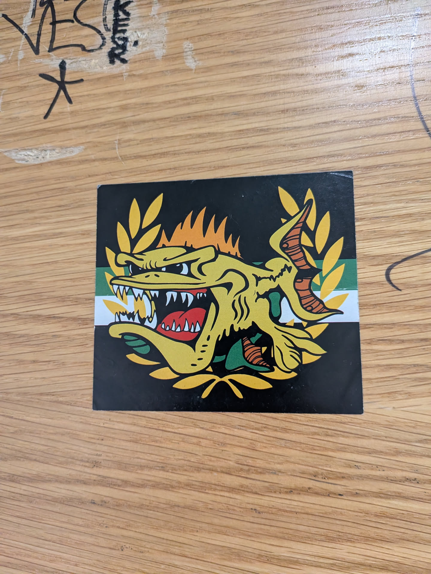 A square sticker featuring a stylized, aggressive yellow fish with sharp teeth and an open mouth. The fish is framed by a yellow laurel wreath against a black background with a horizontal green and white stripe.