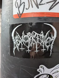 Street sticker A rectangular black sticker featuring a highly stylized, intricate white logo at its center. The logo is rendered in a 'spiky' or 'dripping' font, characteristic of extreme metal genres, making it almost illegible. It is framed on both the left and right sides by prominent white crescent moon or horn-like shapes that curve inwards towards the logo. Below the main logo, there are several smaller, inverted cross-like or spike-like elements. The sticker has a slightly glossy finish, showing some reflections, and appears to be adhered to a dark, possibly metallic, pole.