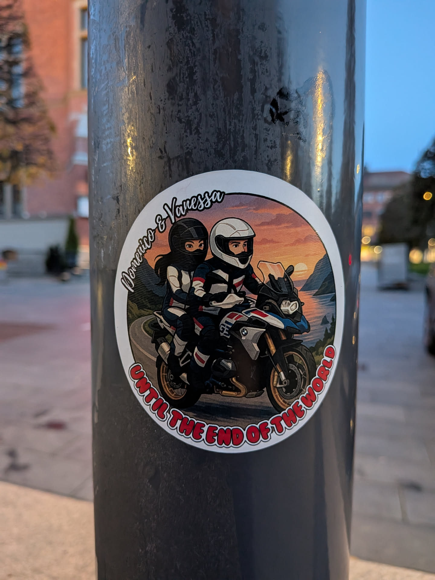 A circular sticker featuring a cartoon illustration of a couple riding a motorcycle. The rider in front has a white helmet and is wearing a black and white riding suit. The passenger behind has a black helmet and is also wearing a black and white riding suit. They are on a white, blue, and black adventure motorcycle. The background depicts a scenic landscape with a winding road, mountains, a body of water, and a vibrant sunset sky with orange, pink, and yellow hues. The sticker has a white border, and text is incorporated into the design.