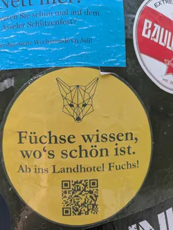 Street sticker The central sticker is circular and yellow with a black border. At the top, there is a black geometric, polygonal illustration of a fox head. Below the illustration, the text "Füchse wissen, wo's schön ist." is written in black. Underneath that, it says "Ab ins Landhotel Fuchs!". At the very bottom, a black QR code is printed on the yellow background.