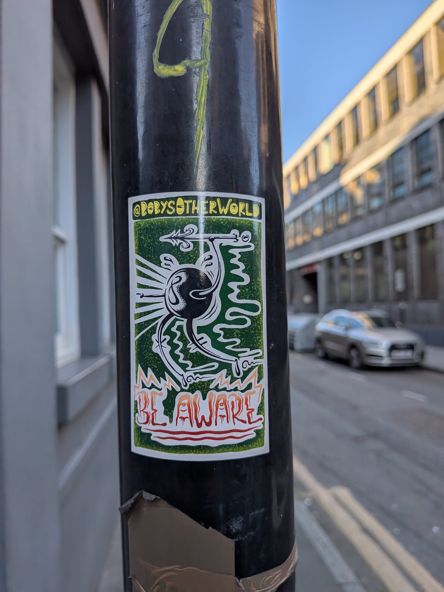 A rectangular sticker featuring a stylized, dark, and somewhat monstrous creature against a mottled dark green background. The creature has a prominent, dark, circular body with a white outline, from which several white-outlined, tentacle-like limbs extend. One limb holds a spear or arrow with a barbed tip, pointing upwards. White rays emanate from the creature's central body. At the top, a yellow banner contains the text "@BOBYSOTHERWORLD" in black. At the bottom, the words "BE AWARE" are written in a dripping, blood-red and orange font with a white outline, set against a jagged white shape.
